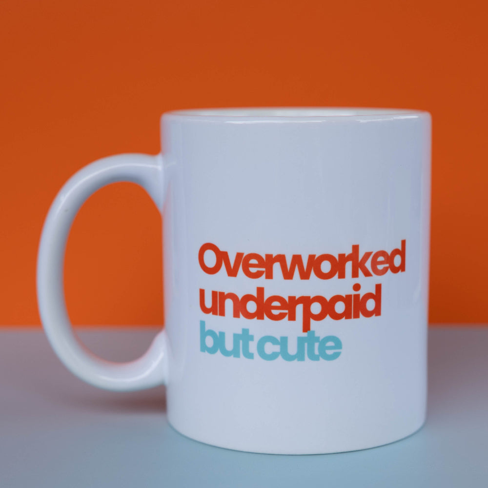 A white ceramic coffee mug with the text 'Overworked underpaid but cute' printed on the side in orange and light blue, set against a split orange and gray background.