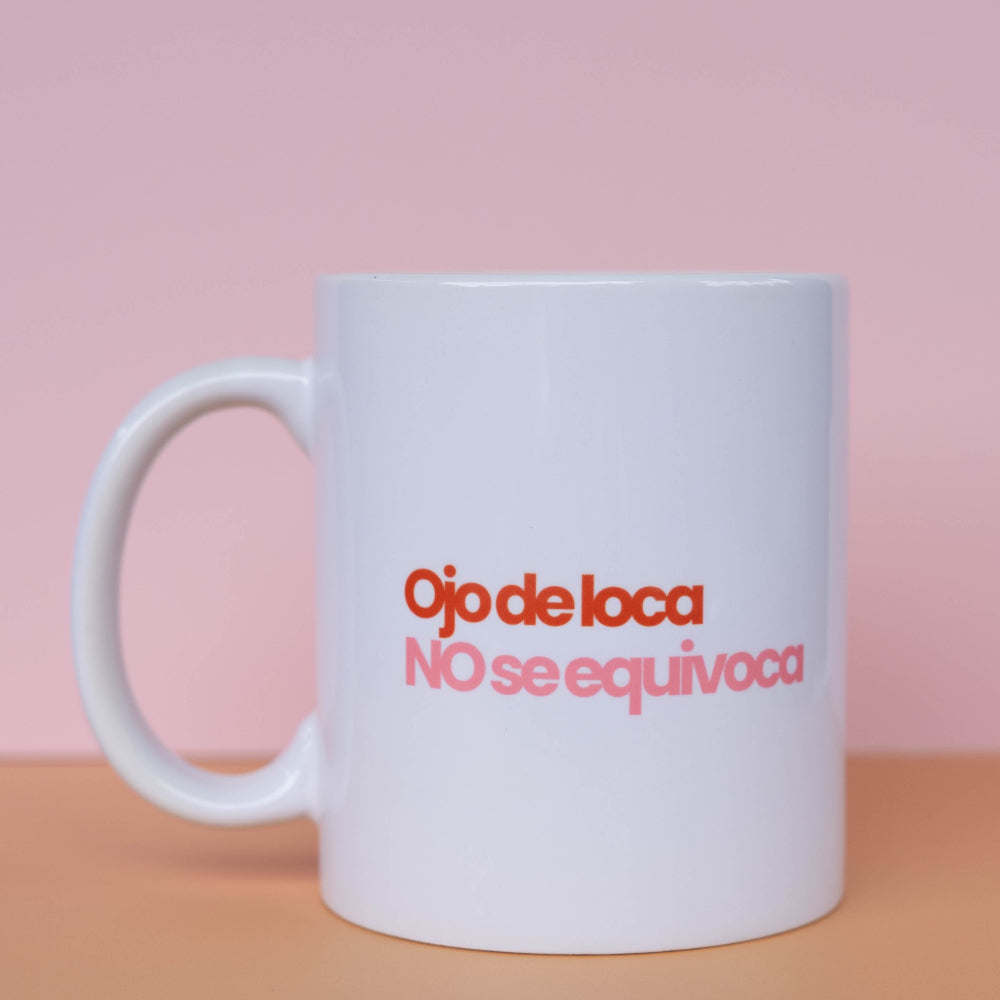 A white ceramic coffee mug with the Spanish text 'Ojo de loca NO se equivoca' printed in red and pink lettering, set against a soft pink background and a peach-colored surface.