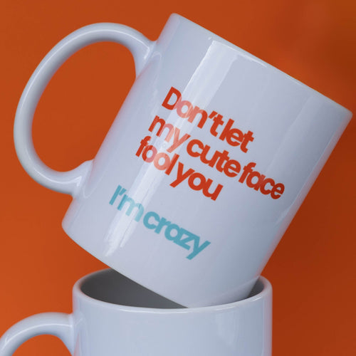 Two white ceramic mugs stacked with a retro typographic design. The top mug is tilted, showing the text 'Don't let my cute face fool you' in orange and 'I'm crazy' in light blue.