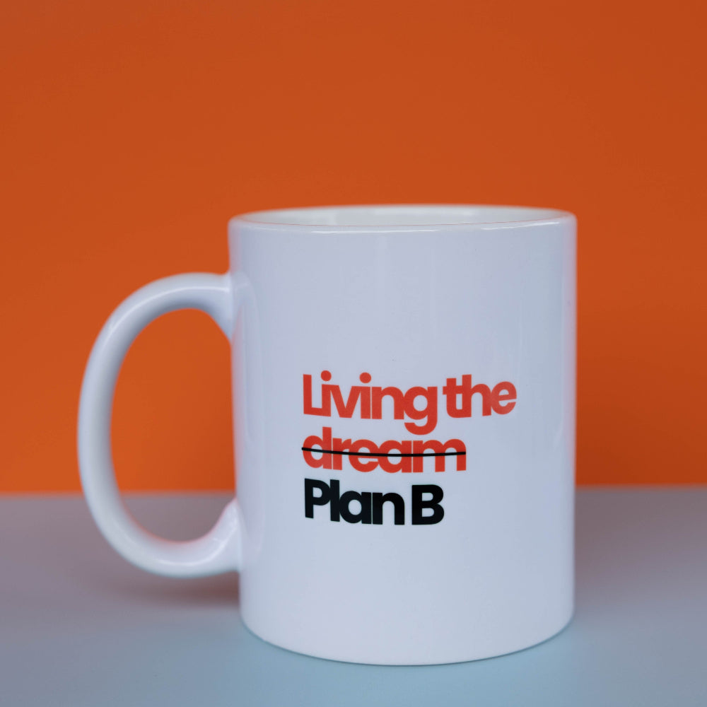 A white ceramic coffee mug with the text 'Living the dream Plan B' printed on the side, set against a vibrant orange background and a light gray surface.