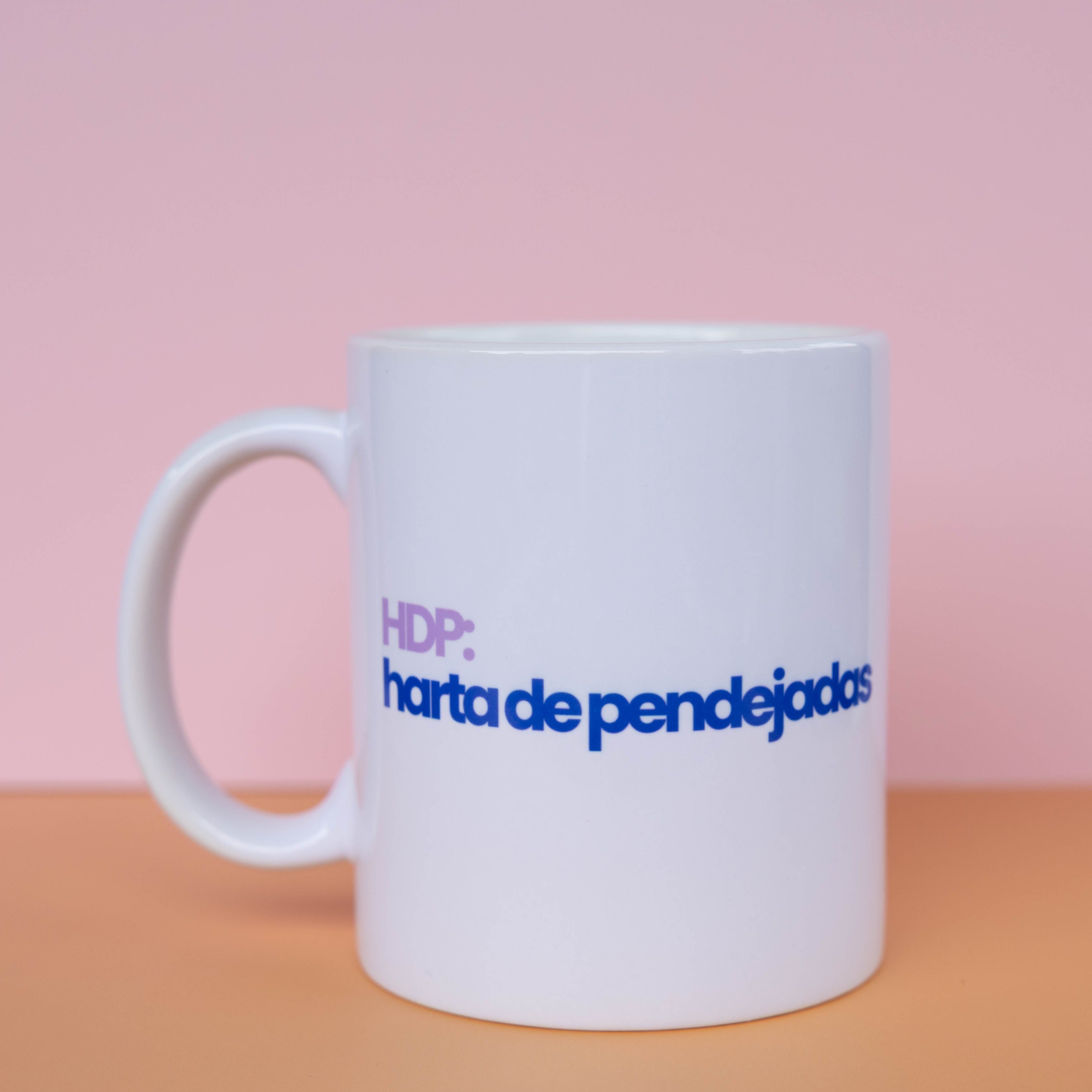 A white ceramic coffee mug with the text 'HDP: carta de pendejos' printed on the side in purple and blue, set against a pink and peach background.