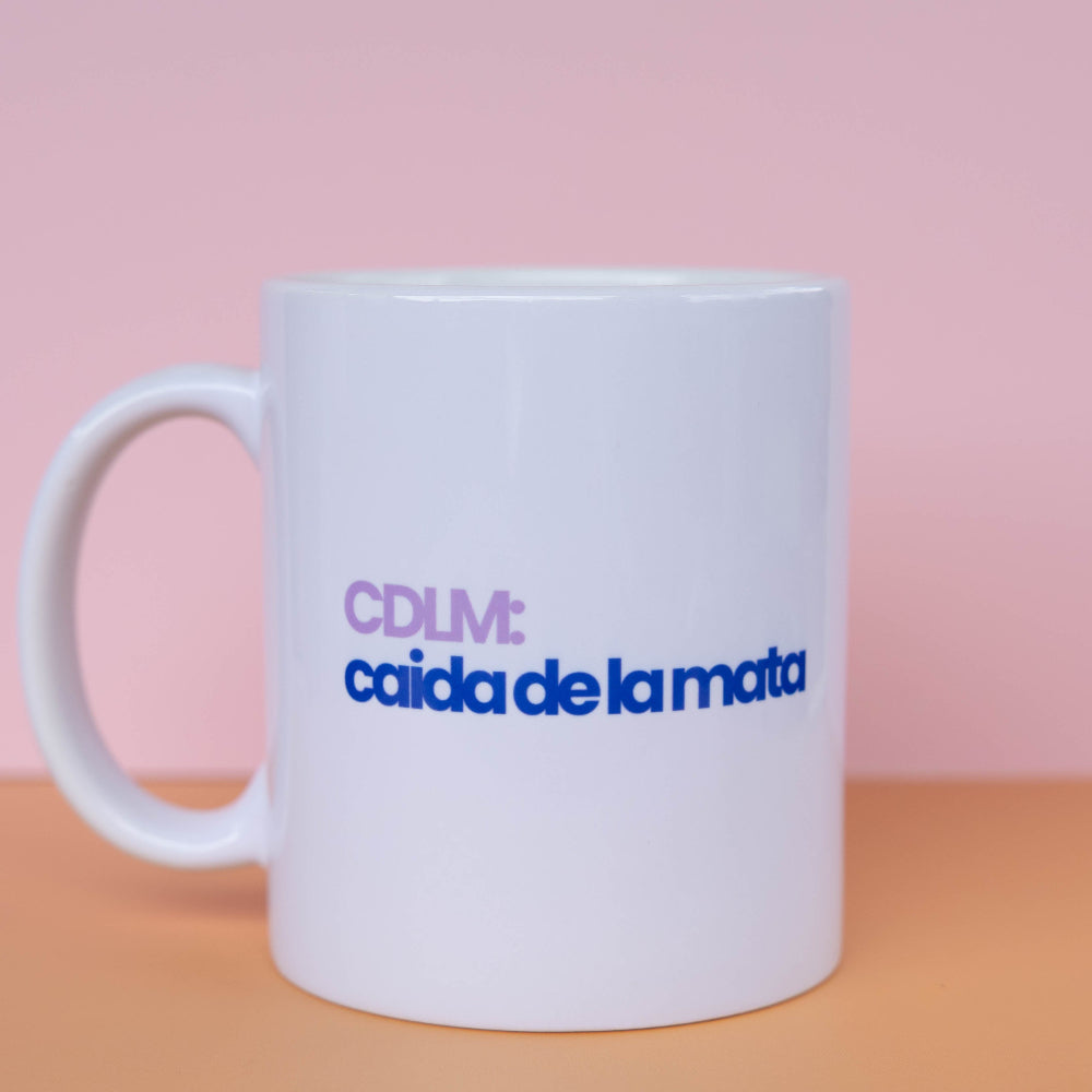 A white ceramic coffee mug with the text 'CDLM: caida de la matra' printed on the side in purple and blue, set against a pink and orange background.