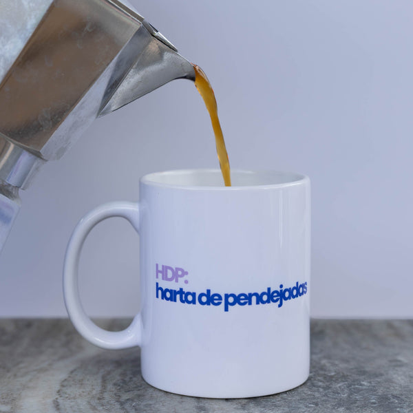 A white ceramic mug with blue and purple text reading 'HDP: harta de pendejados' sits on a gray surface while coffee is being poured from a silver moka pot into the mug by a hand.
