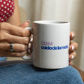 A person holding a white ceramic mug with blue text that reads 'CDLM: caida de la mata' on their lap. The person is wearing a red top with white polka dots, blue jeans, and silver bracelets.