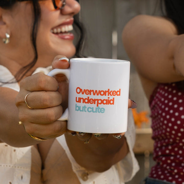 Two women smiling and holding up white mugs with the text 'Overmyrdized underneath' in blue and orange lettering, set against a backyard fence and green leaves.