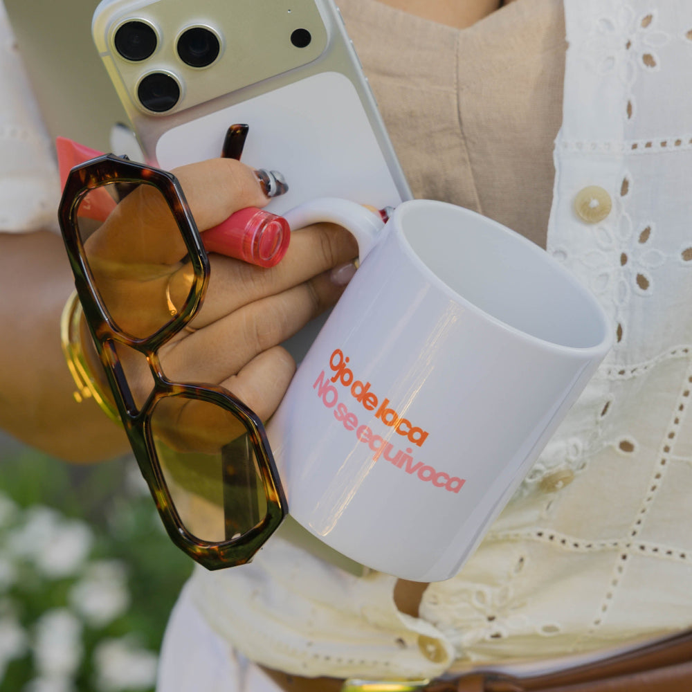 A person outdoors holding a white coffee mug with orange text, a white smartphone in a clear case, sunglasses, and a gold phone case.