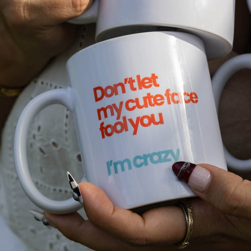 A person in a white eyelet blouse holds four white ceramic mugs printed with colorful Spanish phrases such as 'Blessed, stressed' and 'Overworked underpaid but cute.'.