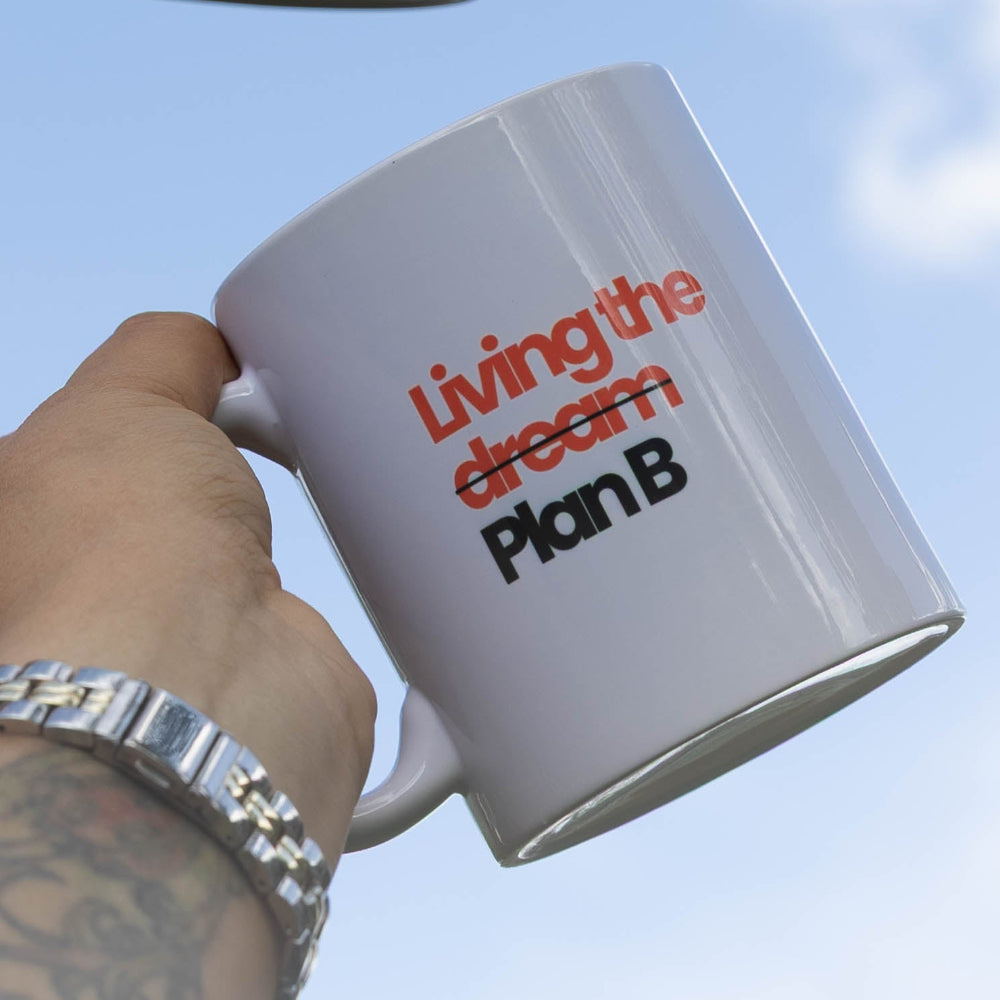 Two hands holding up white ceramic mugs with Spanish text against a blue sky with clouds. One mug reads ' Blessed, unpressed y sin tiempo para que vivan' and the other reads 'living the design PlanB'.