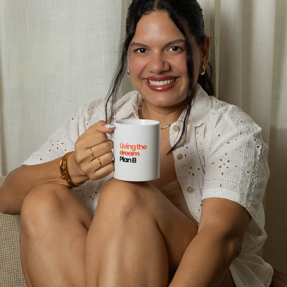 A woman sits cross-legged in a beige armchair holding a white mug with a red logo. She is wearing a white short-sleeve top and white knit Mary Jane-style shoes.