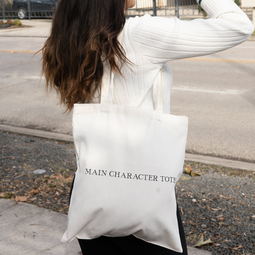 A woman outdoors wearing a gray baseball cap, white ribbed sweater, and black pants, carrying a large off-white tote bag printed with the text 'MAIN CHARACTER TOTE'.